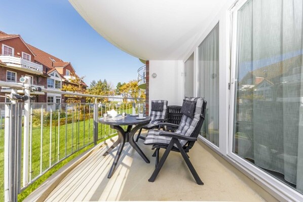 Un balcon avec une table et deux chaises, une porte-fenêtre coulissante et une vue sur un quartier résidentiel avec des maisons et des arbres.