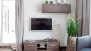 Een moderne woonkamer met een flatscreen televisie aan de muur, een houten kastje eronder en een potplant aan de rechterkant.
