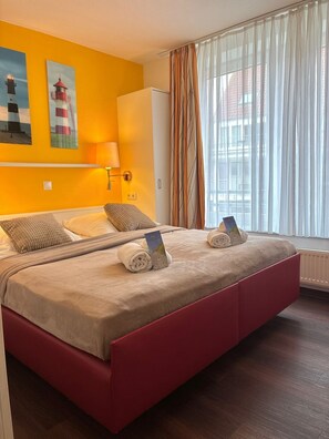 Uma cama de casal com estrutura vermelha, cabeceira branca e colcha bege. Há dois travesseiros, uma luminária e uma janela com cortinas leves.
