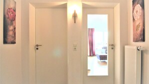 Een gang met twee deuren, waarvan er een leidt naar een kamer met een radiator en een raam met gordijnen.