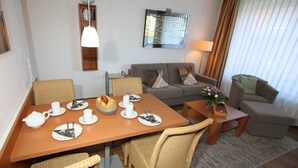A dining area with a wooden table set for four, surrounded by wicker chairs. A sofa and a small side table with a vase of flowers are in the background.