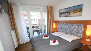 A hotel room with a large bed, a wooden headboard, a painting on the wall, a balcony with a table and chairs, and a view of buildings outside.