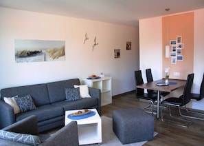 A modern living room with a grey sofa, a small white side table, a dining area with a wooden table and black chairs, and a wall with framed pictures and a bird sculpture. - HLI 3.24 4 people 48sqm - Residenz Hohe Lith Cuxhaven (3.2) (Cuxhaven)