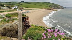 A coastal landscape with a sandy beach, a stone wall, and a white building with a car parked in front. - Beach Front Coastal Retreat with Sea Views (Kingsbridge)