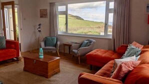 A living room with a large window, a red sofa, a grey armchair, a wooden coffee table, and a tripod. - Beach Front Coastal Retreat with Sea Views (Kingsbridge)