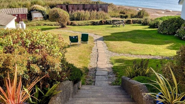 A coastal property with a grassy lawn, a stone staircase leading to a deck, and a view of the sea. - Beach Front Coastal Retreat with Sea Views (Kingsbridge)