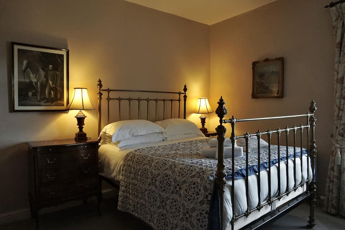 A bedroom with a four-poster bed, two bedside tables with lamps, a nightstand, and two framed pictures on the walls.