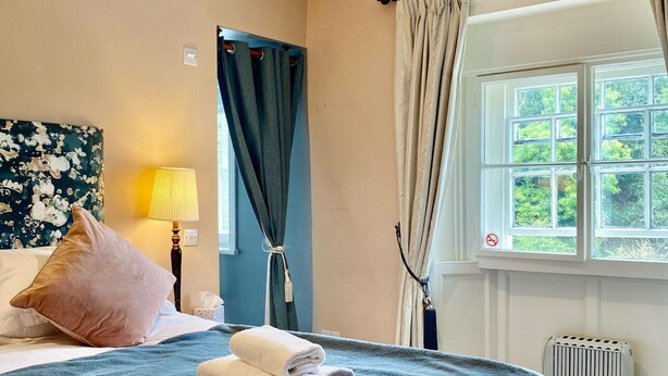 A hotel room with a bed, a window with curtains, a radiator, and a wall with a floral painting. - The Derby Arms Witherslack (Grange-over-Sands)