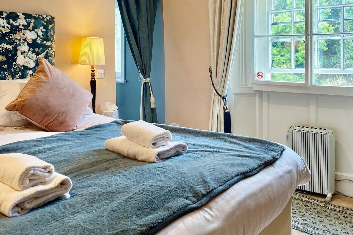 A hotel room with a bed, a window with curtains, a radiator, and a wall with a floral painting.