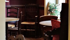 A dining area with wooden chairs and a table, a red wall with framed portraits, and a window with a patterned curtain. - The Derby Arms Witherslack (Grange-over-Sands)