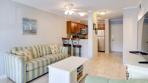 A living room with a striped sofa, a white coffee table, and a green armchair. There is a kitchen area with wooden cabinets and a refrigerator. - Beachy Getaway w/ Balcony on Hilton Head Island! (Hilton Head Island)