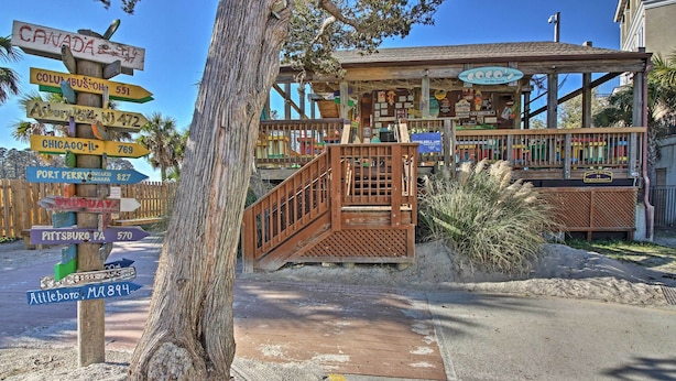 A wooden signpost with multiple directional signs, a wooden staircase leading to a building with a sign reading "Coco's", and a sandy ground area. - Beachy Getaway w/ Balcony on Hilton Head Island! (Hilton Head Island)