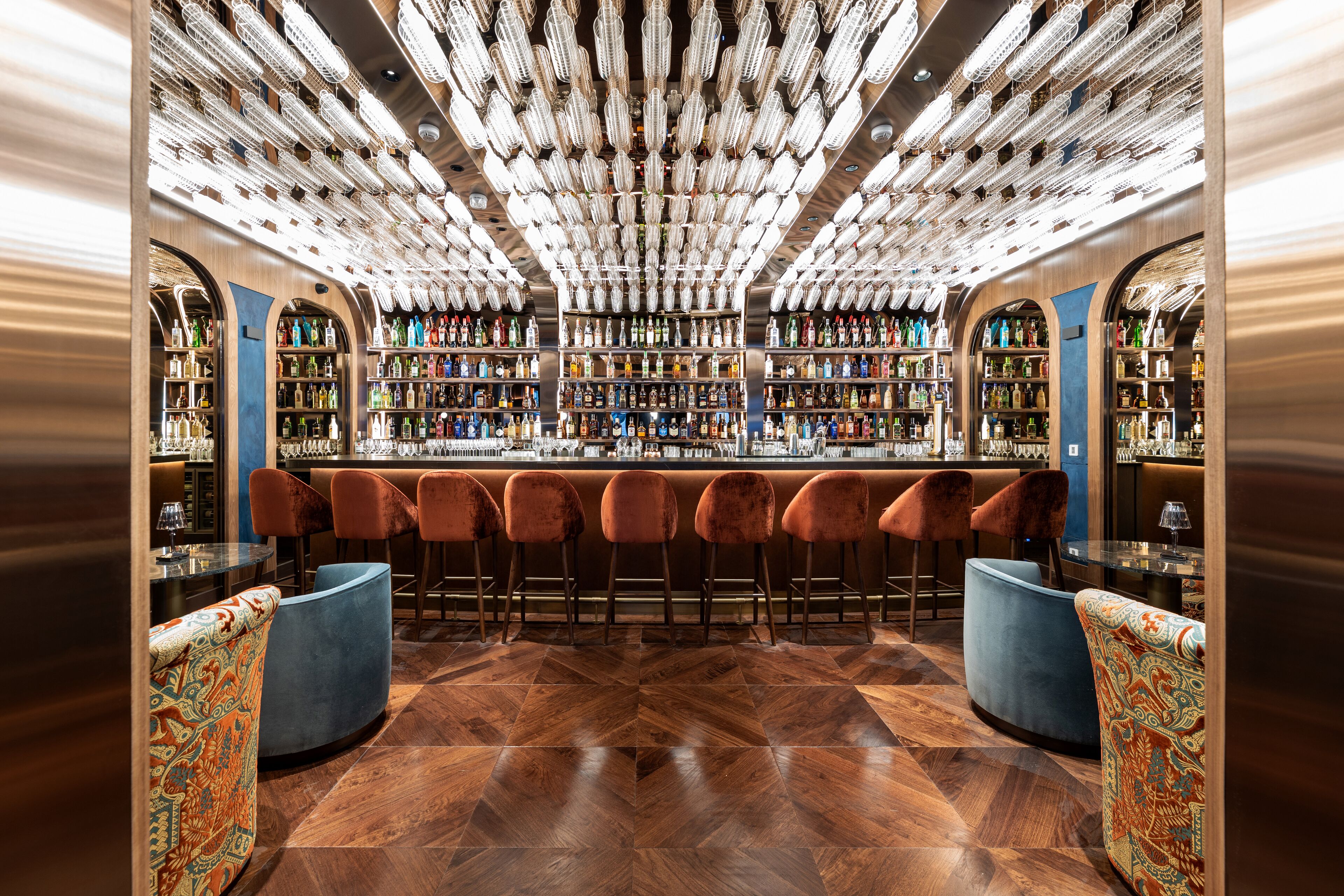 A bar area with a wooden herringbone floor, velvet bar stools, and a bar counter with a variety of bottles.