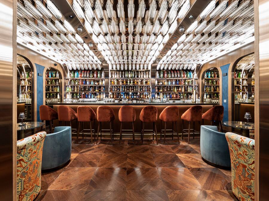 A bar area with a wooden herringbone floor, velvet bar stools, and a bar counter with a variety of bottles.