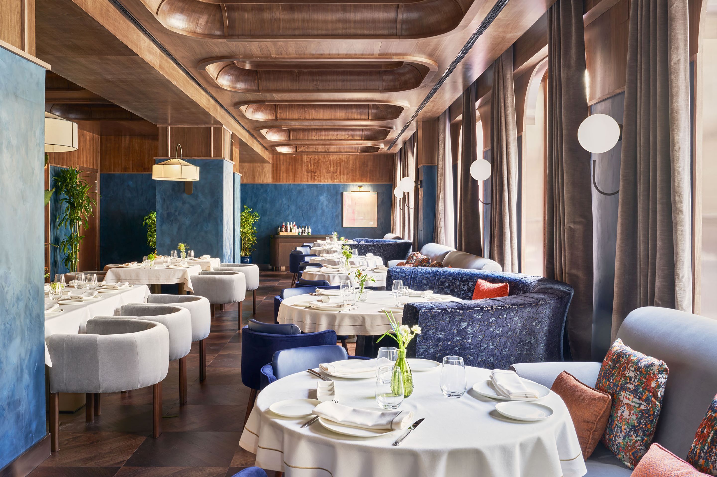 Um restaurante com mesas redondas e assentos estofados em azul, com teto de madeira e grandes janelas.