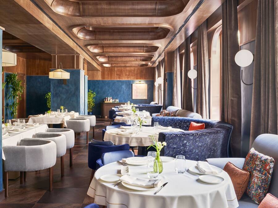 Um restaurante com mesas redondas e assentos estofados em azul, com teto de madeira e grandes janelas.