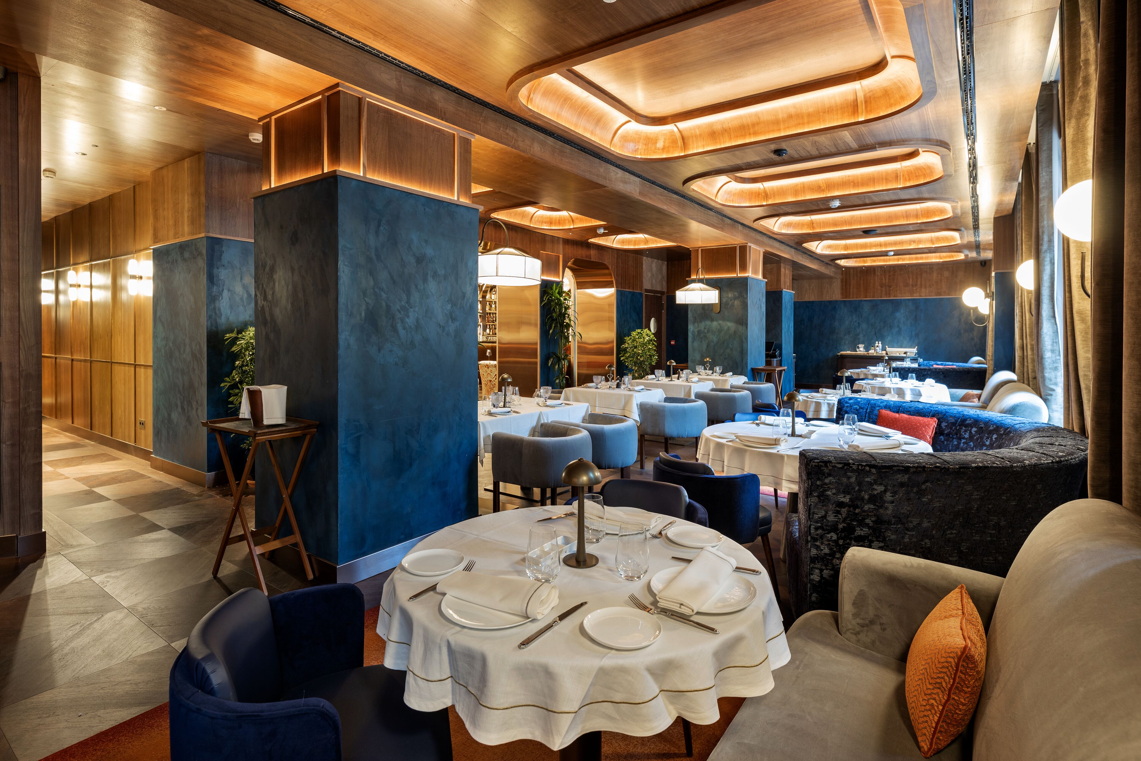 Un comedor bien iluminado con mesas redondas dispuestas para una comida, sillas tapizadas en azul y un diseño moderno en el techo.