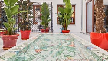 A spacious indoor area with a tiled floor featuring a colorful mosaic pattern, potted plants, and a building with large windows and balconies.