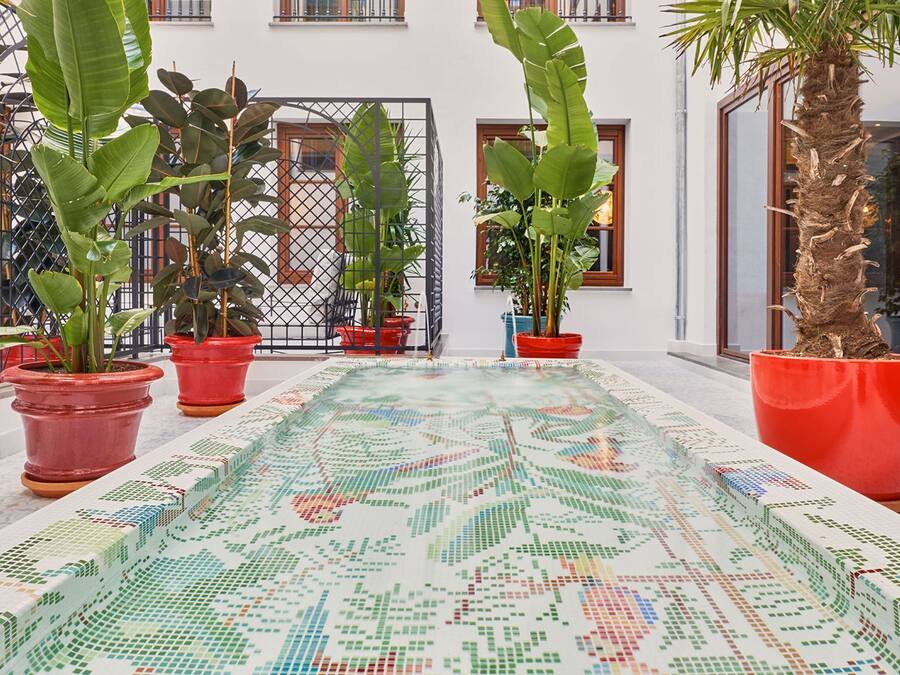 A spacious indoor area with a tiled floor featuring a colorful mosaic pattern, potted plants, and a building with large windows and balconies.