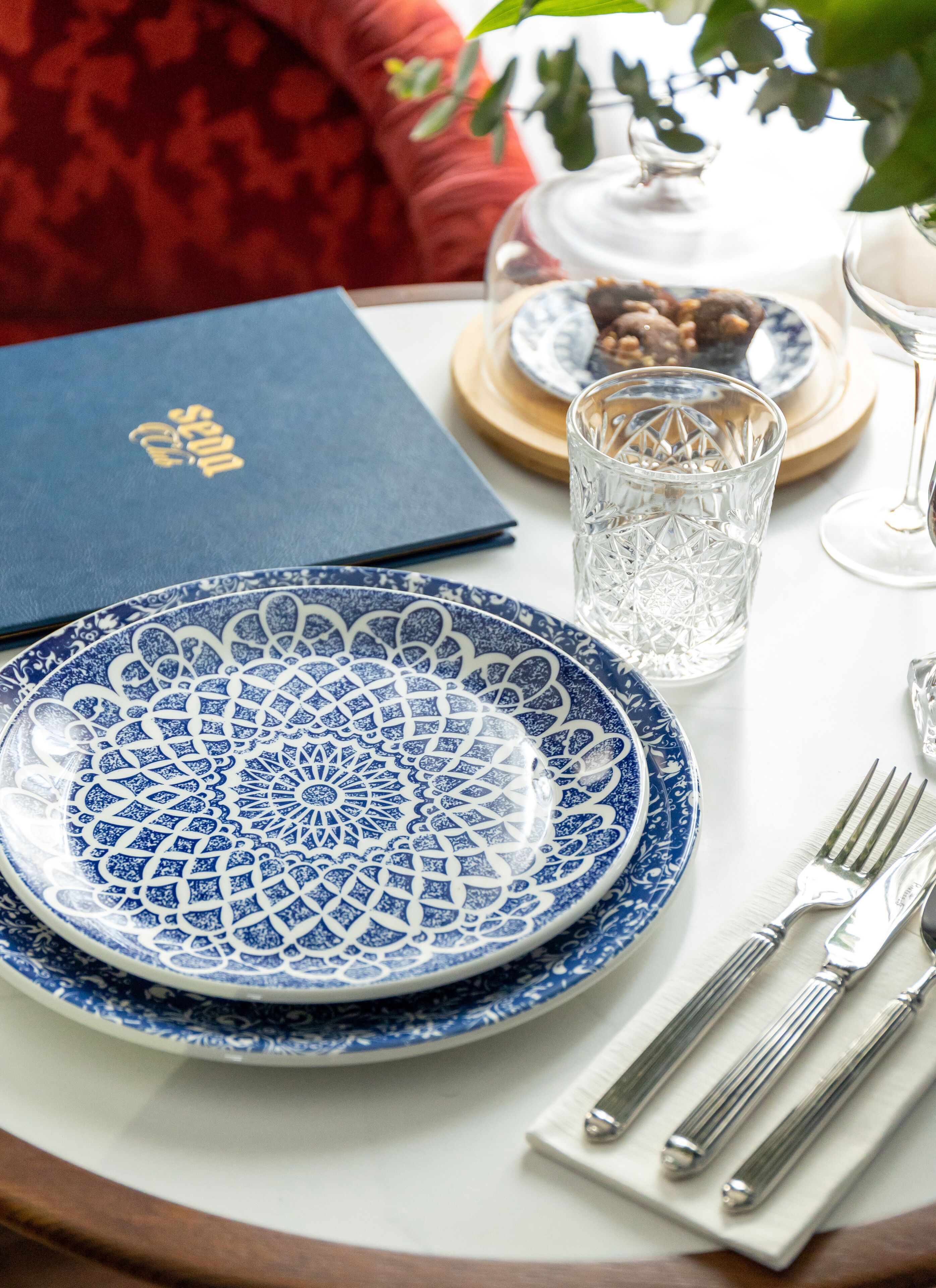 Una mesa dispuesta con un plato azul y blanco estampado, una copa de cristal transparente, un menú azul y cubiertos sobre un mantel blanco.