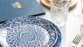 Una mesa dispuesta con un plato azul y blanco estampado, una copa de cristal transparente, un menú azul y cubiertos sobre un mantel blanco.