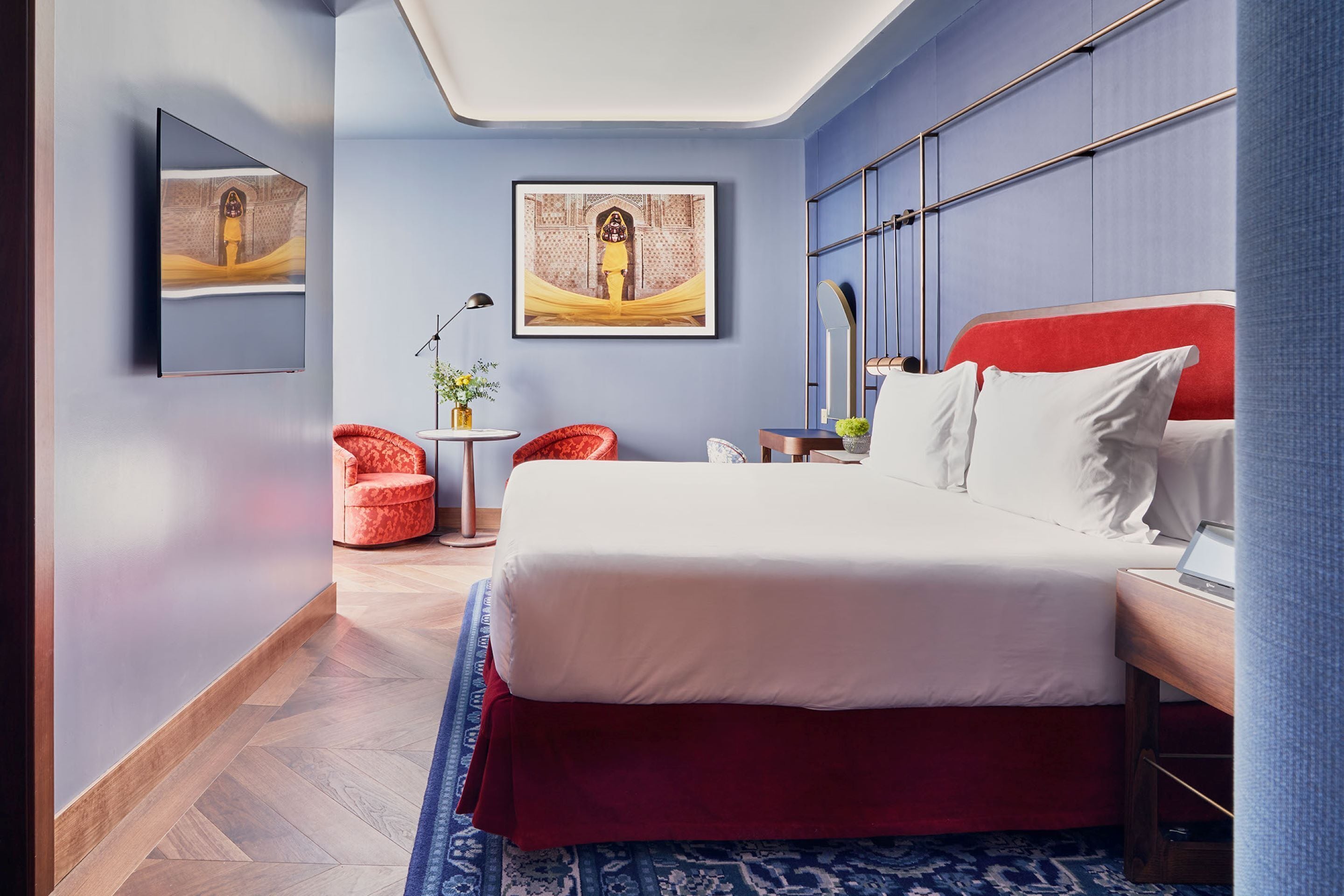 A modern hotel room with a large bed, a red headboard, and a blue accent wall. There is a small table with a lamp and a plant, and a red chair.