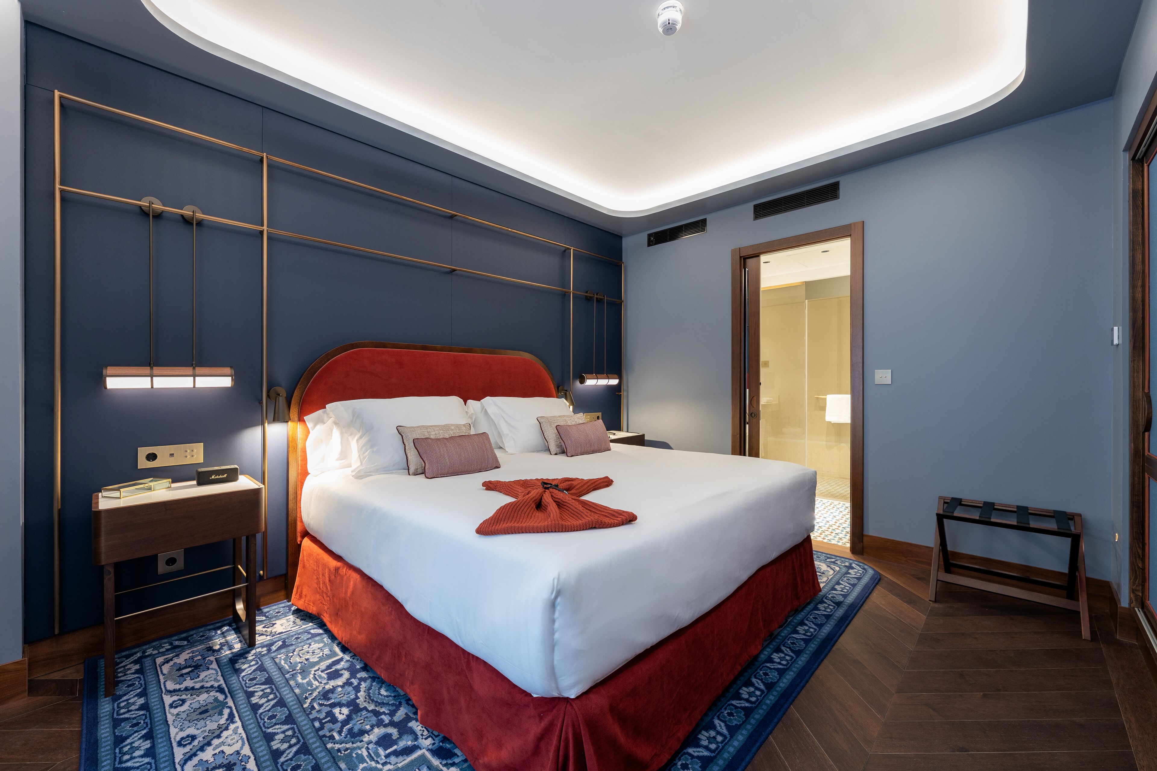 Moderni hotellihuone, jossa on suuri sänky, yöpöydät, yövalaisin, pieni penkki ja erillinen vaatehuone.