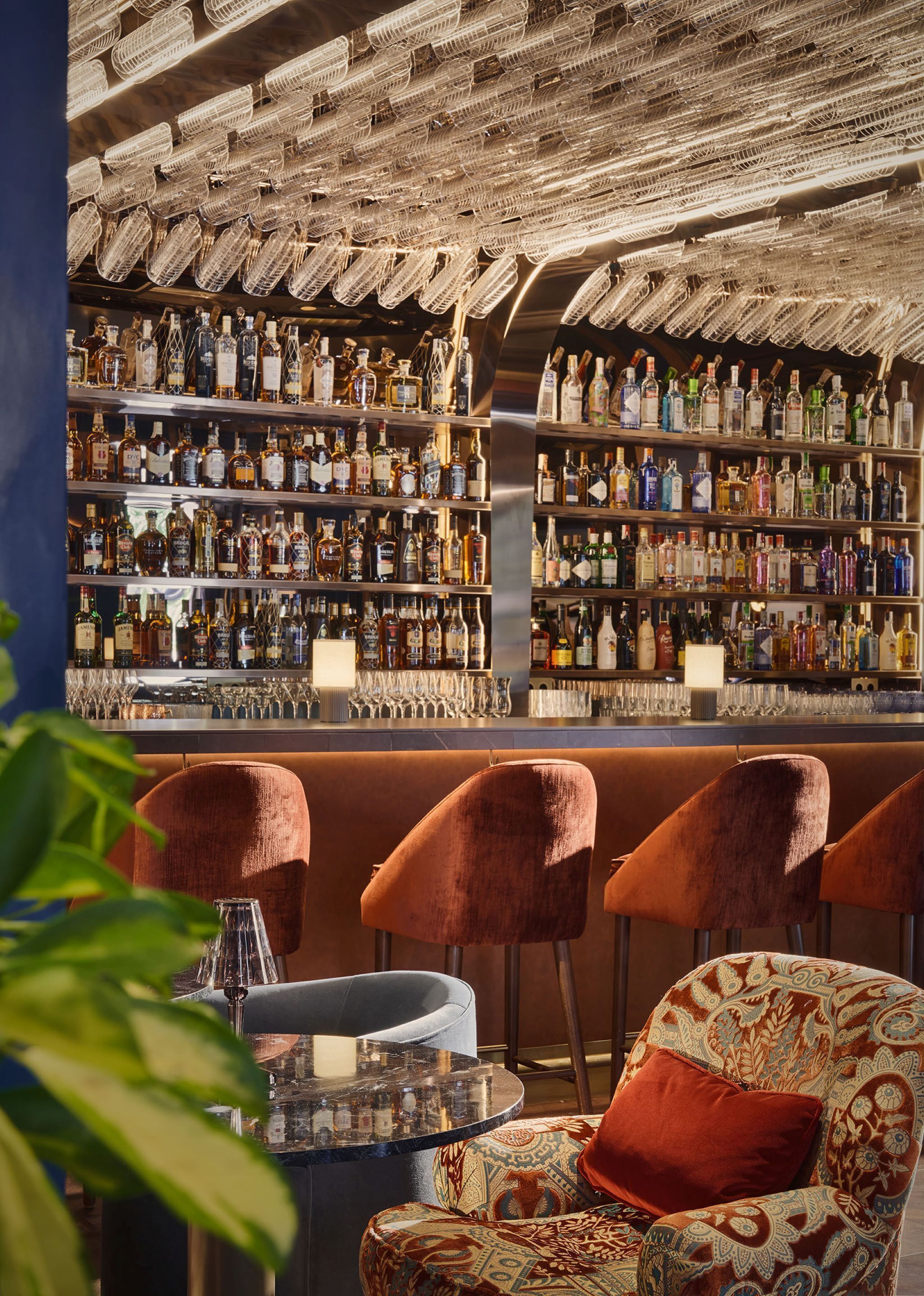 Zona de bar con estantes llenos de botellas de licor, barra y asientos con tapicería estampada.