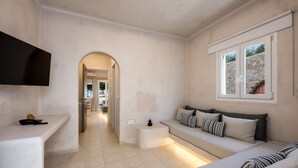 A modern hotel room with a sofa, a TV, a small table, and a window with a stone wall view. - Aelia Luxury Suites (Santorini)