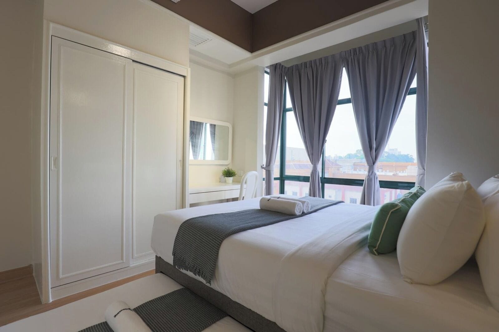Ett hotellrum med en säng, en garderob, ett fönster med gardiner, ett litet bord med en växt och utsikt över byggnader utanför.