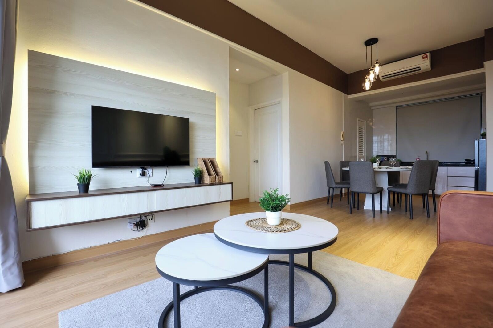 Una sala moderna con un televisor de pantalla plana montado en la pared, dos mesas redondas con sillas, un comedor y una cocina al fondo, y un sofá marrón.