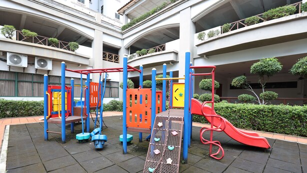 A playground with climbing structures and a slide, surrounded by a building with multiple balconies and greenery.