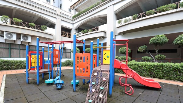 Ein Spielplatz mit Klettergerüsten und einer Rutsche, umgeben von einem Gebäude mit mehreren Balkonen und Grünflächen.