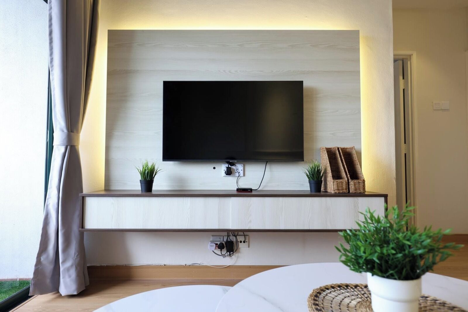 Una sala moderna con una televisión de pantalla plana montada en una pared de madera, un mueble bajo blanco con dos plantas en macetas y una cesta de mimbre.
