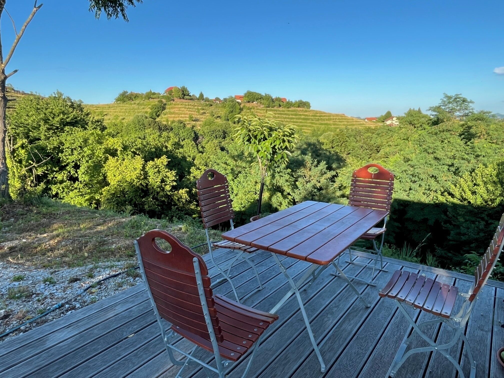 A wooden deck with a table and chairs, overlooking a lush green landscape with hills and trees.