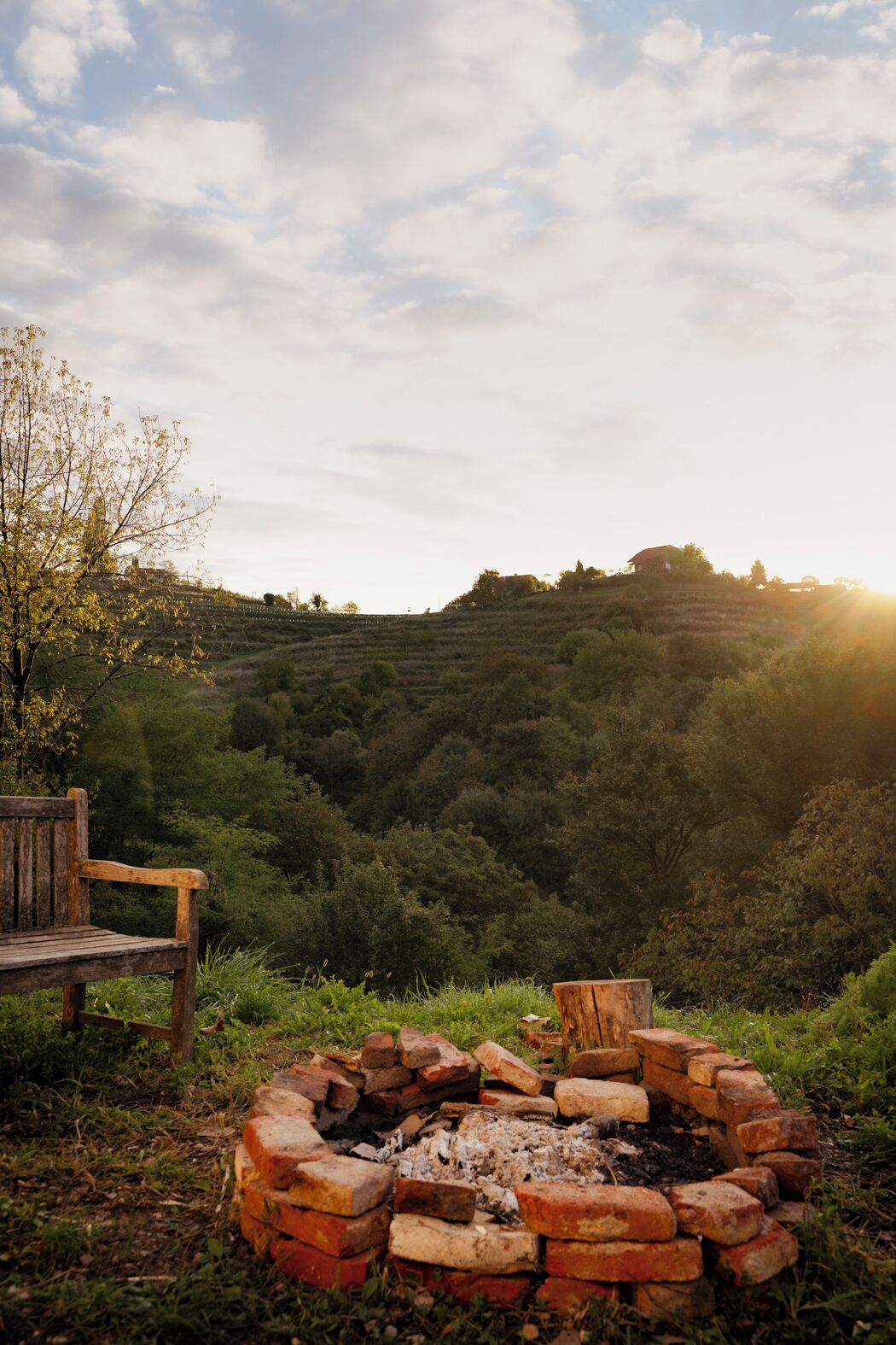 Un'ambientazione esterna rustica con una panchina di legno, un braciere in mattoni e una collina sullo sfondo.