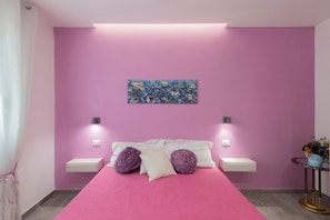 Un dormitorio con pared rosa, una cama con cubrecama rosa, dos almohadas moradas, una mesita de noche blanca, una pequeña mesa redonda con una planta y una obra de arte en la pared.