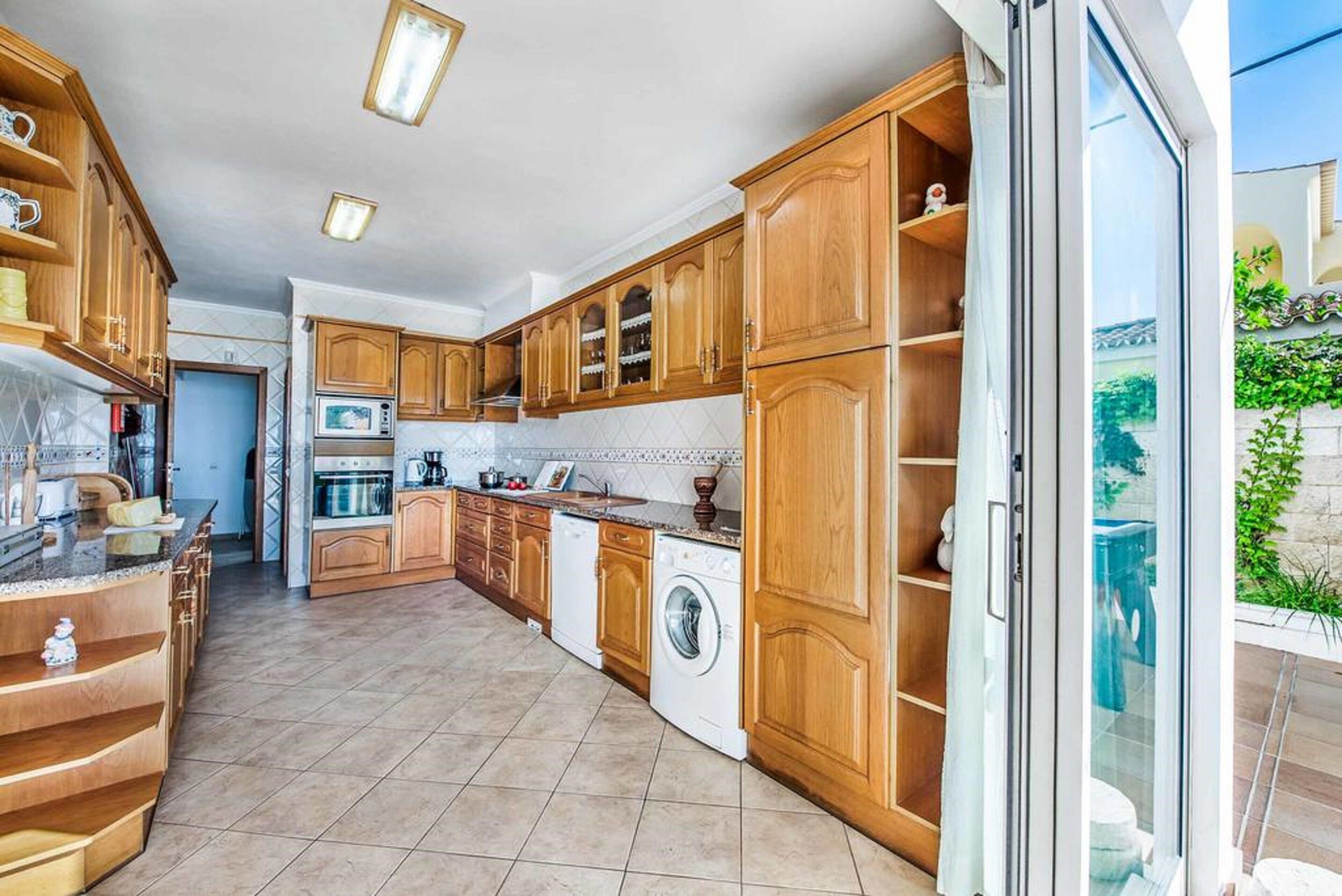 A modern kitchen with wooden cabinets, a built-in oven, a washing machine, and a large window.