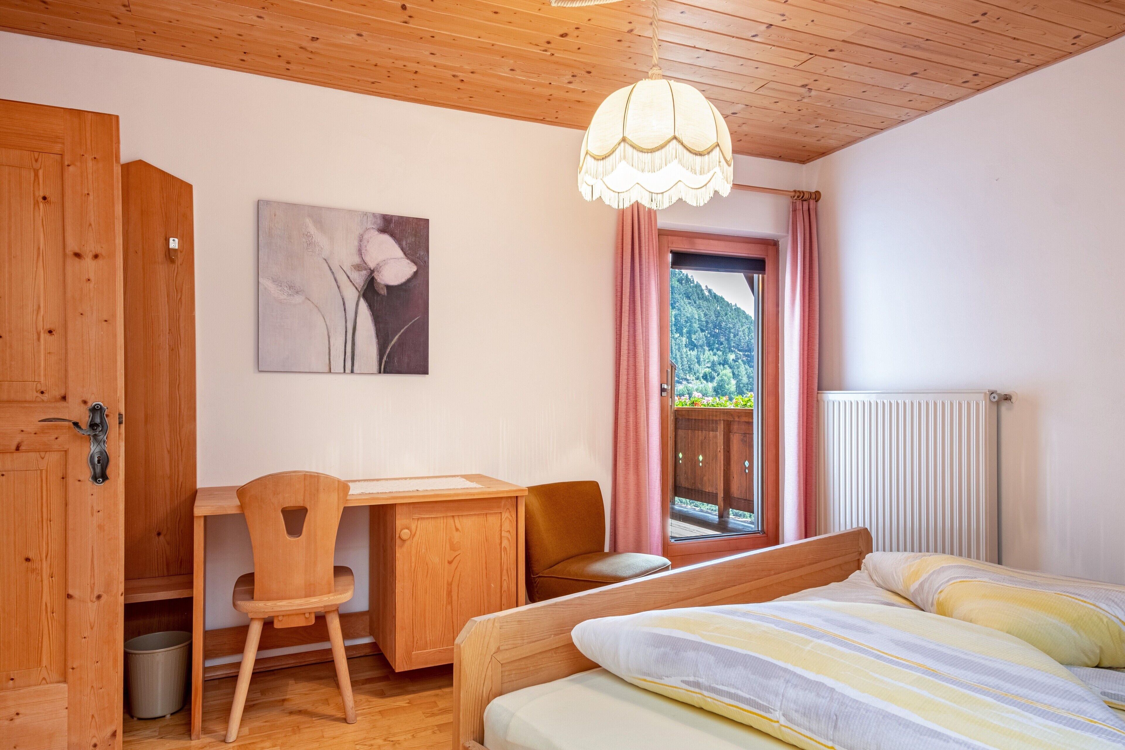 A room with a wooden desk, chair, and bed, a radiator, and a window with a view of greenery.