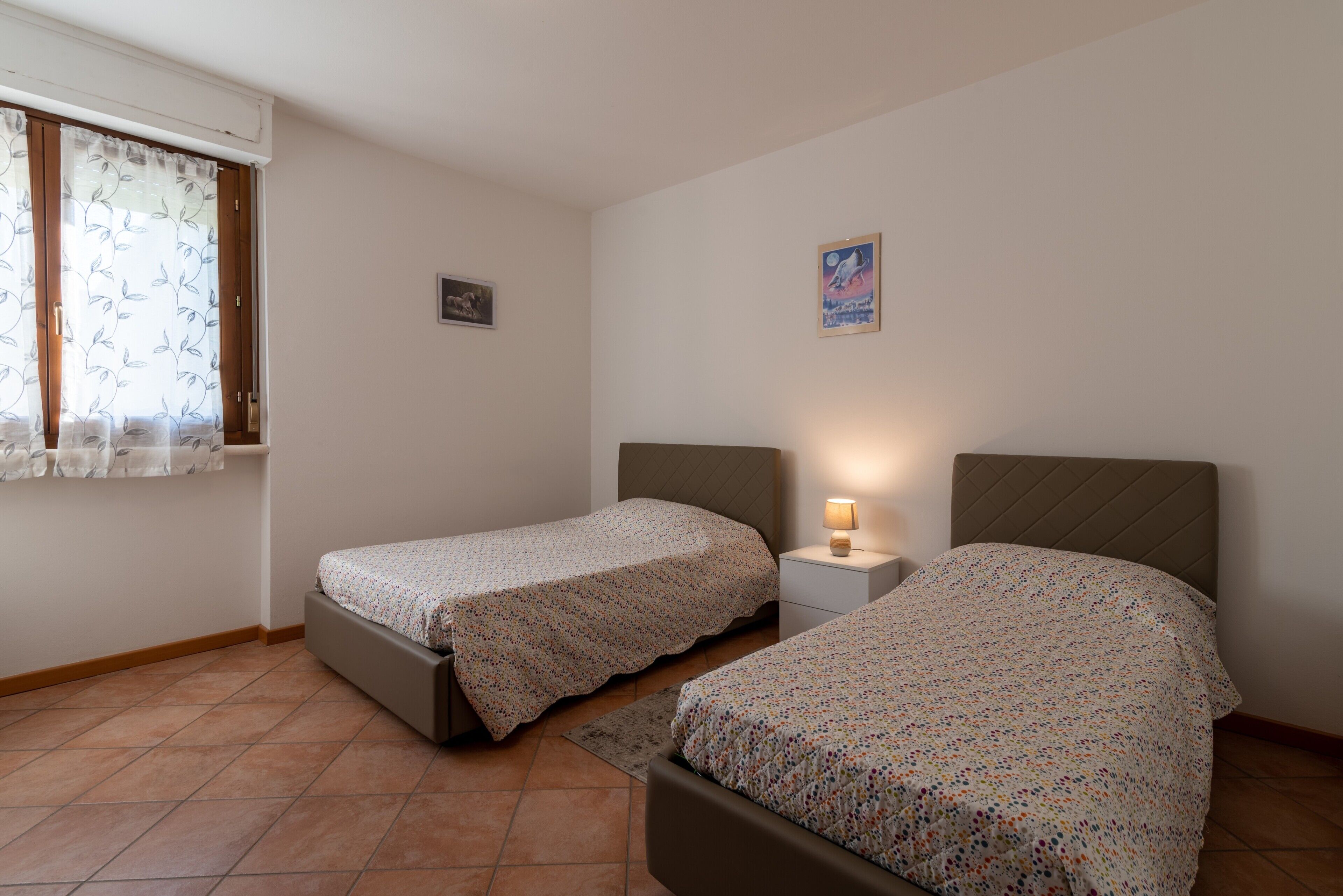 Quarto com duas camas de solteiro, mesa de cabeceira com luminária, janela com cortinas e quadro fixado na parede.