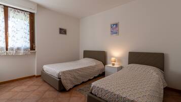 Quarto com duas camas de solteiro, mesa de cabeceira com luminária, janela com cortinas e quadro fixado na parede.