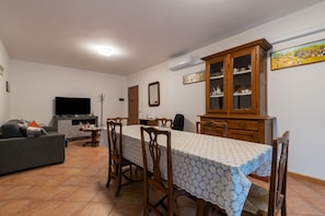 Ein Essbereich mit einem Tisch, gedeckt für vier Personen, umgeben von Holzstühlen. Im Hintergrund befindet sich ein Fernseher und ein Schrank mit Glastüren.
