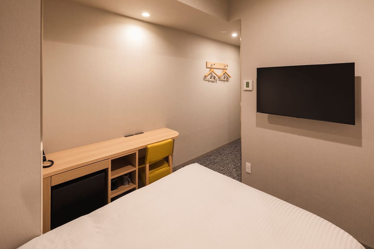 Habitación de hotel con cama, escritorio con silla, televisor montado en la pared y estante pequeño con perchas.