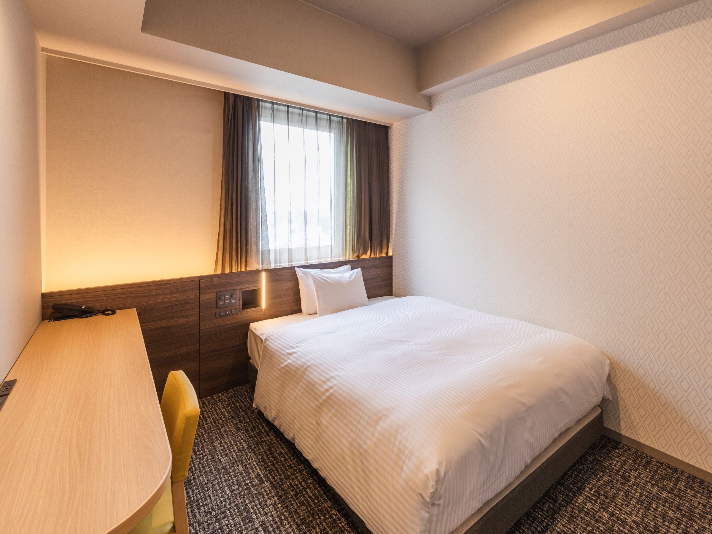 A hotel room with a bed, a desk, a chair, a window with curtains, and a wall with a geometric pattern.