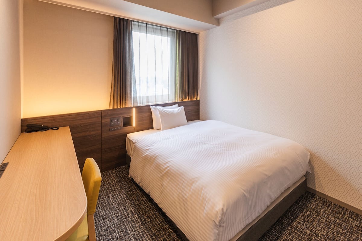 Habitación de hotel con cama, escritorio, silla, ventana con cortinas y pared con diseño geométrico.