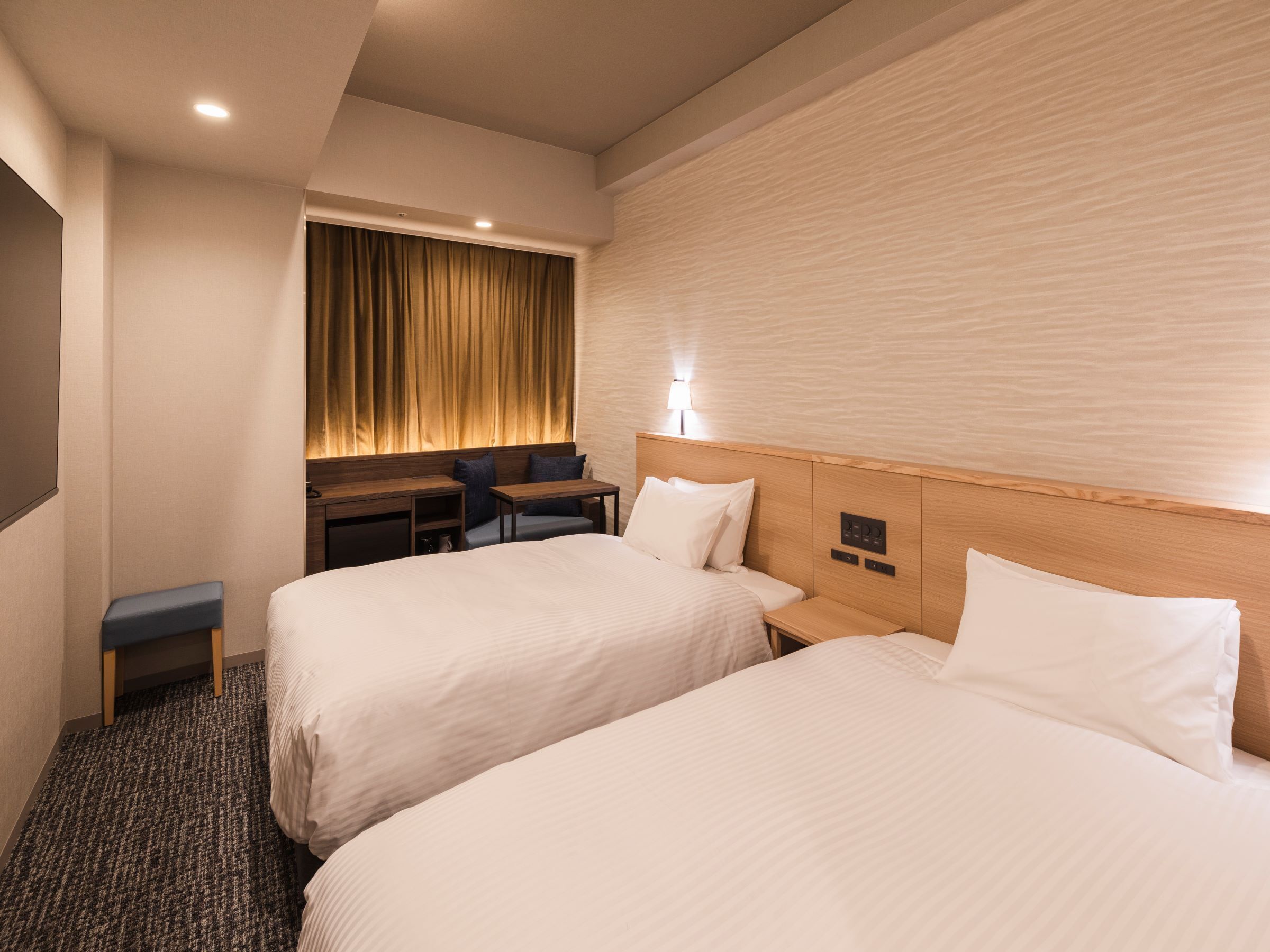 A hotel room with two beds, a desk, a chair, and a wall-mounted lamp.