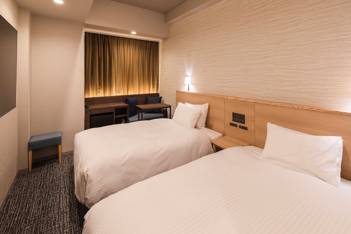 Una habitación de hotel con dos camas, un escritorio, una silla y una lámpara fijada a la pared.