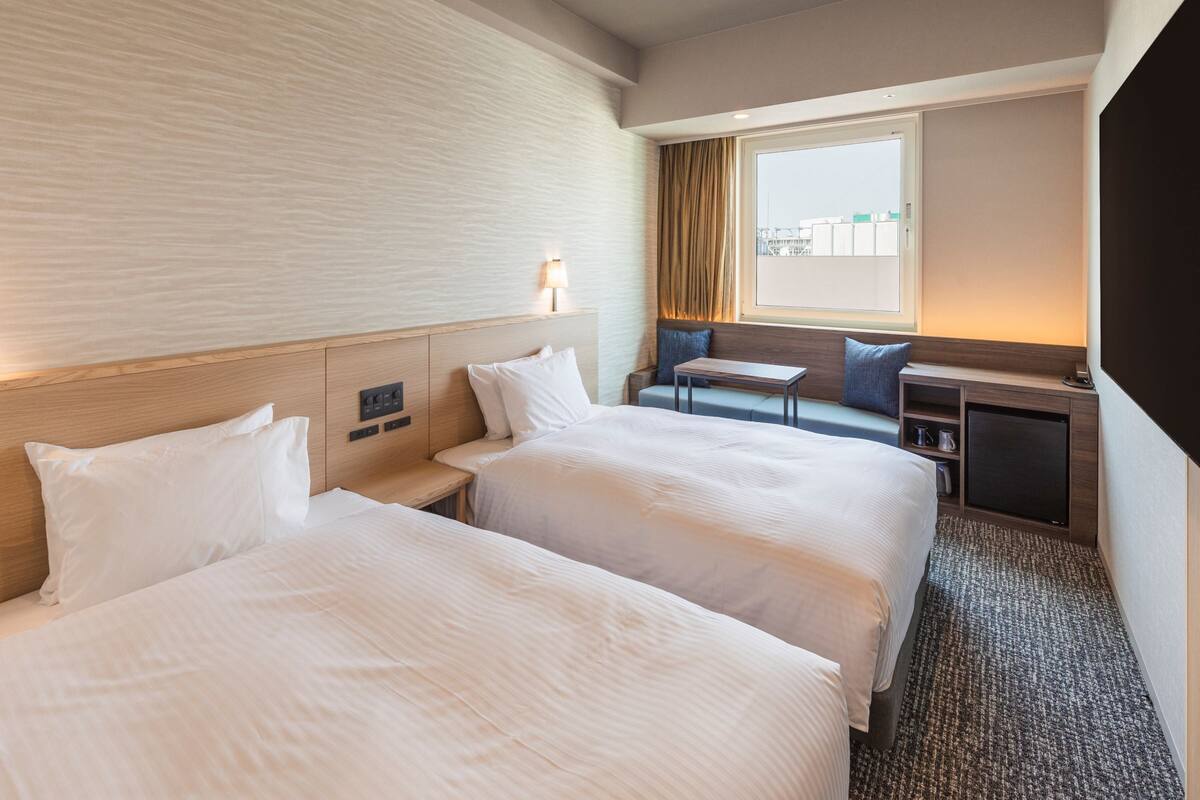 Una habitación de hotel con dos camas, un escritorio, una silla, un ventanal con cortinas y un televisor.