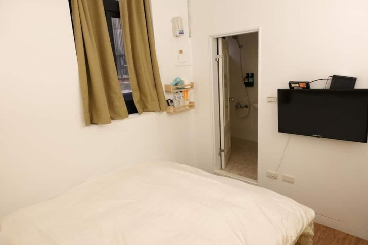 Une chambre à coucher comprenant un lit, un téléviseur fixé au mur, une porte donnant sur une salle de bain et une fenêtre avec des rideaux.