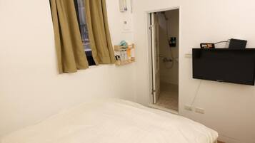 A bedroom with a bed, a TV mounted on the wall, a door leading to a bathroom, and a window with curtains.
